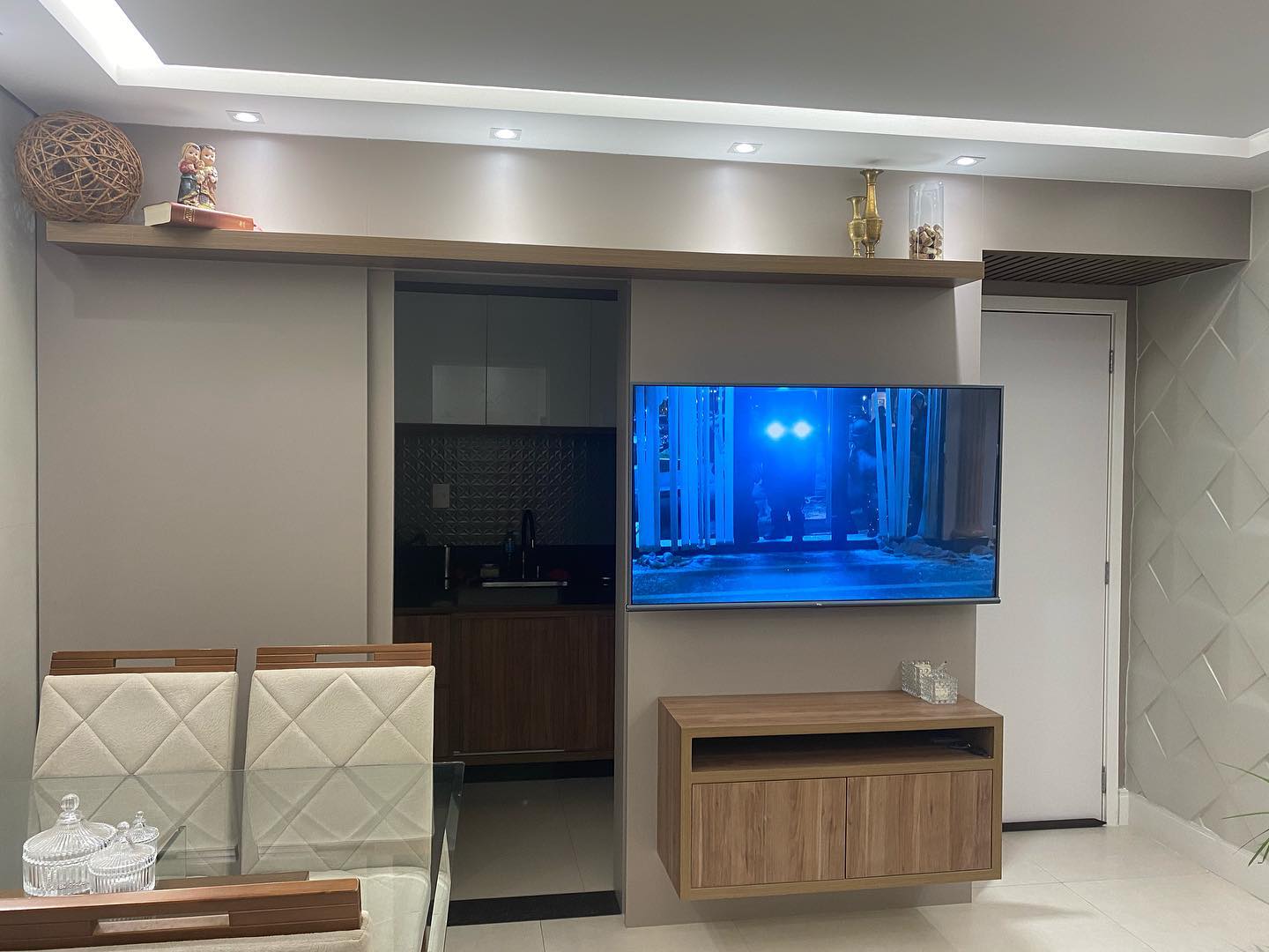 Sala integrada com painel de madeira, iluminação embutida e passagem para a cozinha